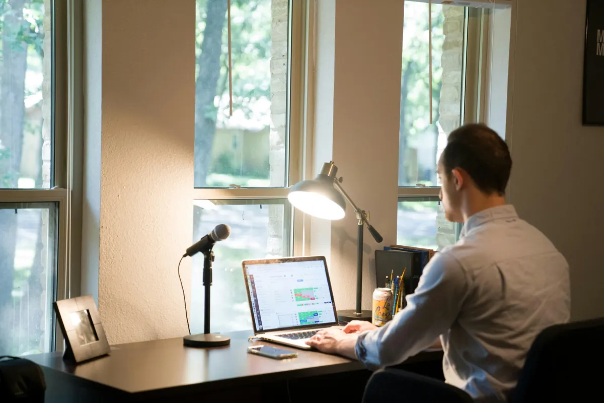 Man working on a laptop at a desk by the window, with a microphone and a desk lamp