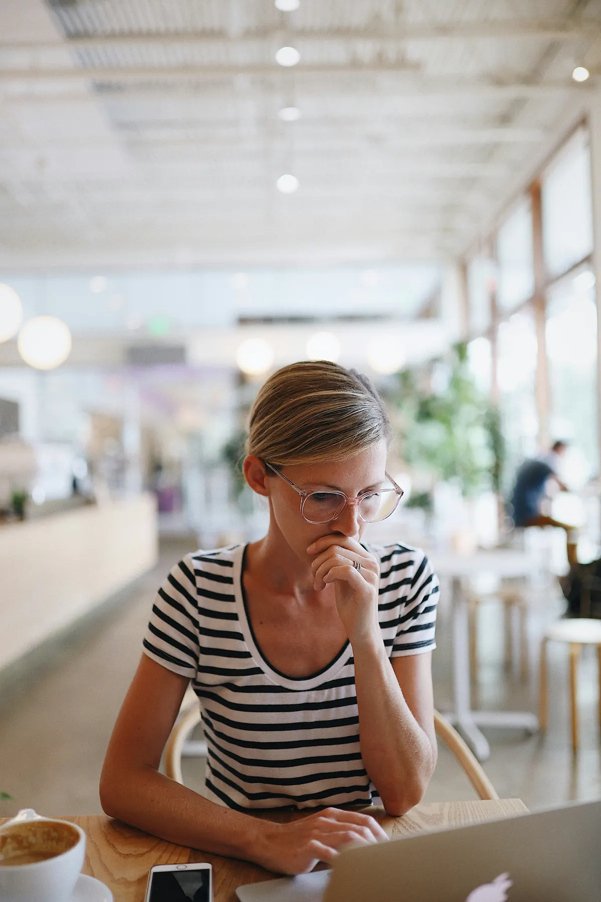 Woman at a laptop in a cafe, thinking with her hand at her mouth